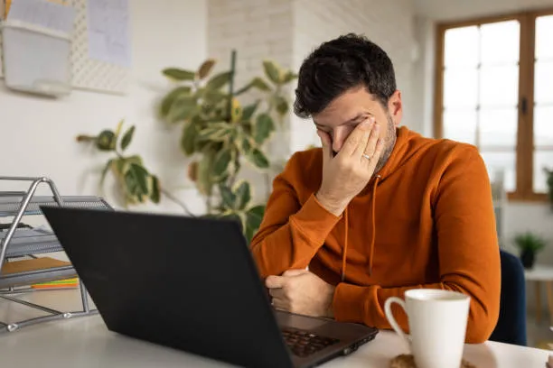 Man covering face while working on laptop, showing stress, fatigue, or burnout
