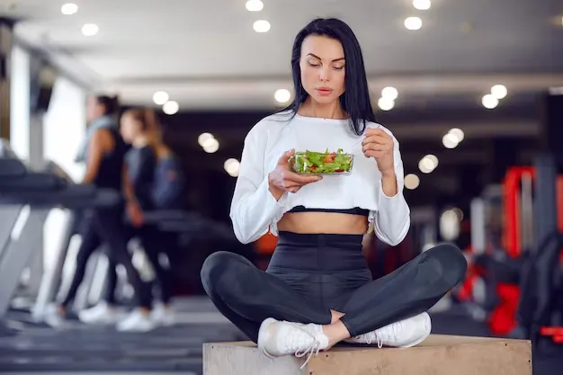 Woman sitting cross-legged at gym eating a healthy salad, mindful nutrition
        