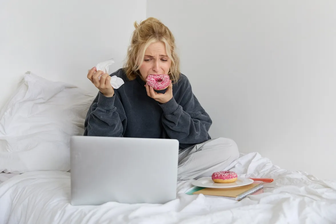 Woman eating donut in bed while using laptop, showing emotional eating or cravings
